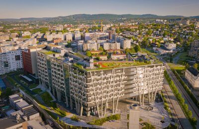 Et stort næringsbygg i fokus med glass og metallfasader. En grønn dag i Oslo med Holmenkollåresen i bakgrunn.