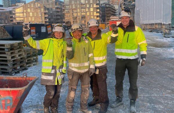 Fire glade mennesker i gule jakker og hvit hjelm foran en byggeplass