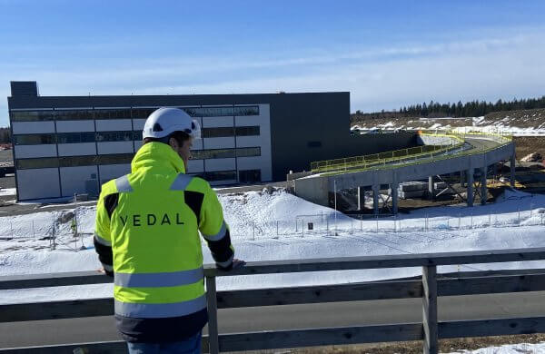 Mann med gul jakke og logoen "Vedal" og hvit hjelm står på en veranda og ser ut på et logistikkbygg