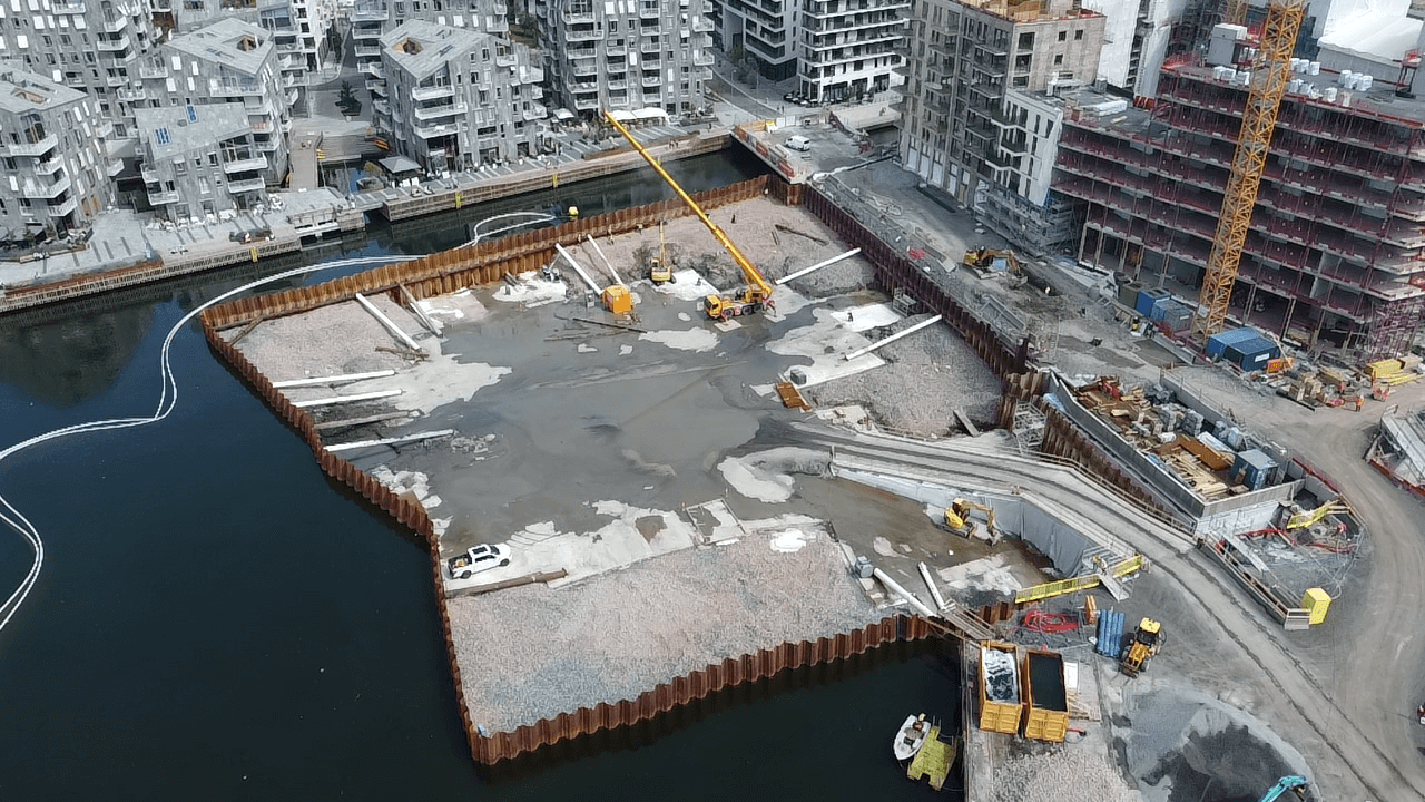 Aktuelt - Byggegrop for Vannkunsten syd boliger. Etablering av byggegrop i sjø. Spuntvegger er etablert i sjøen med lekter. Spuntvegger er stabilisert med støttefyllinger av stein. Byggegrop er videre tømt for vann og sikret med innvendige hvite skråstivere og støpt arbeidsplattform i betong. Fra støpt arbeidsplattform utføres pelefundamentering og betongarbeider for kjelleren.